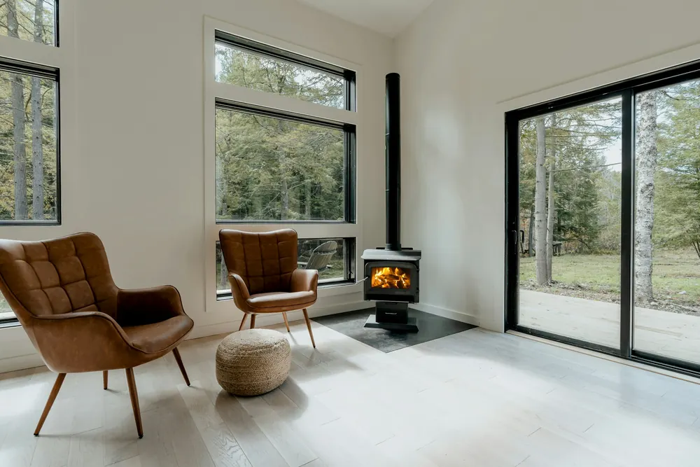 Salon lumineux avec deux fauteuils en cuir face à un poêle à bois allumé près d'une baie vitrée