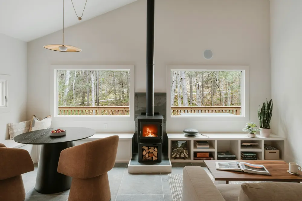 Pièce de vie claire et contemporaine, poêle à bois allumé entre deux fenêtres donnant sur les arbres